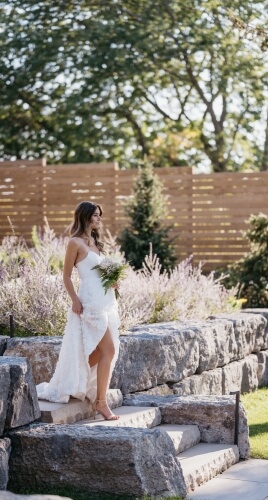 Bride, in a white dress holding a bouquet of white flowers, walks down steps at the Lakecroft ceremony site surrounded by purple lilacs